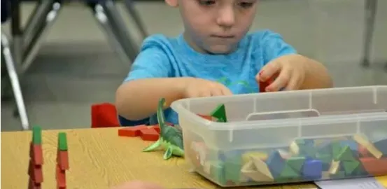 young boy sorting blocks at school desk with plastic bin of building blocks in front of him