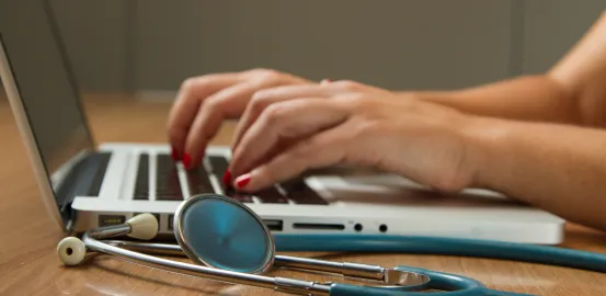two hands typing on the keyboard of a laptop; stethoscope laying next to laptop