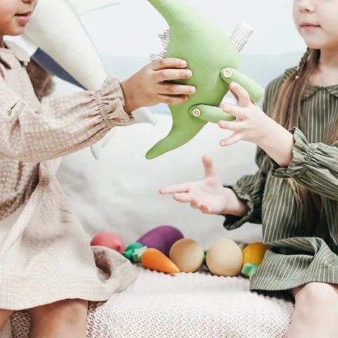 two young girls taking turns playing with a green dinosaur toy