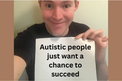 A man looks at the camera and holds a sign that says "Autistic people just want a chance to succeed" 