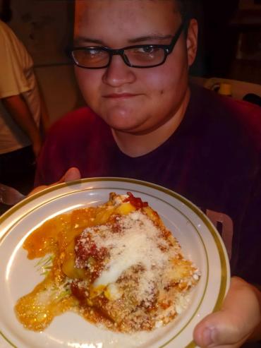 A young man in glasses wears a red shirt and has lasagna in front of him 