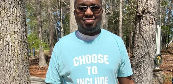 Young black man with glasses on stands in a teal t-shirt that says "Choose to Include" in white lettering 