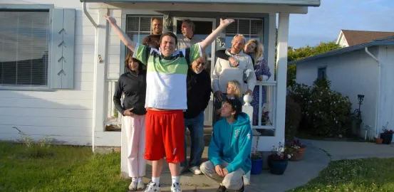 A young man stands in front of his new home, surrounded by his family, friends, and caregivers