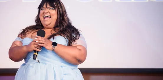 Aarti Garg laughing and holding a microphone while wearing a light blue off the shoulder dress at a public speaking event