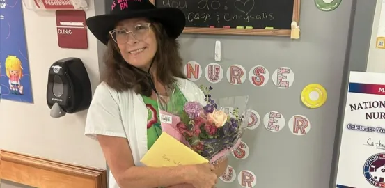 Cathey, RN, in a nurse's coat and cowboy hat, holding flowers in a decorated hallway.