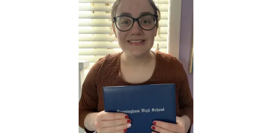 A girl with black glasses and a maroon shirt holds a blue high school diploma to the camera and is smiling 