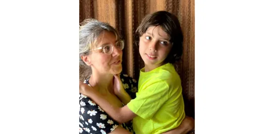 A young boy in a  bright yellow shirt looks out in the distance in his mother's arms, as the mother in glasses and a polka dot shirt looks at him 