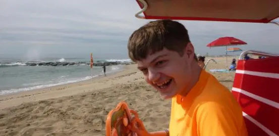 A young boy in an orange shirt sits on the beach under a red umbrella. He is holding a device and smiling at the camera 