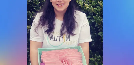 A women in a white shirt that has a AutismWish logo on it holds a teal fabric box with pink letters