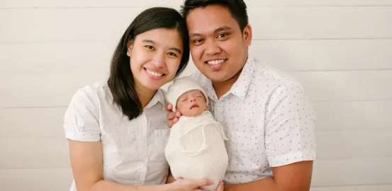 Smiling couple holding a newborn baby wrapped in white blanket against a light background