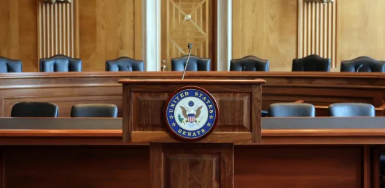 Wooden podium with US Senate seal in front of two lines of long committee desks with black chairs