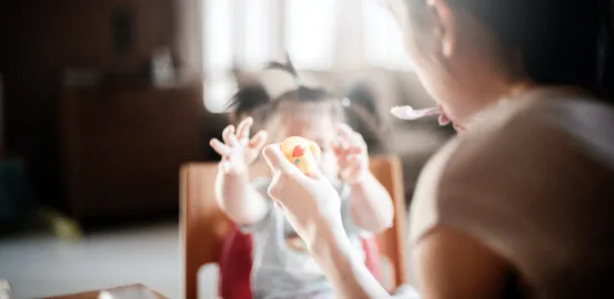 a toddler in a highchair reaching for food as her mother feeds her