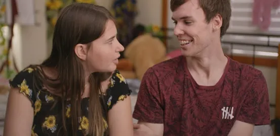 A white couple is smiling at each other mid laugh. The girl has a black shirt with sunflowers on it, and the boy has a red shirt. 