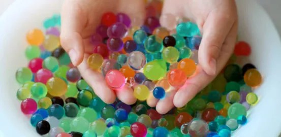 two hands holding a bunch of colorful water beads over a white bowl of more water beads