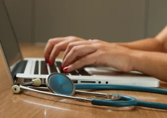 two hands typing on the keyboard of a laptop; stethoscope laying next to laptop