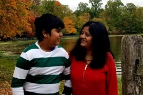 A teenager of Indian heritage  with a green and white striped shirt and looks at his mother standing in a red shirt in front of colorful fall trees 