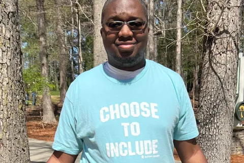 Young black man with glasses on stands in a teal t-shirt that says "Choose to Include" in white lettering 