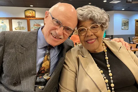 A man with glasses putting his arm around a woman with glasses as they both smile