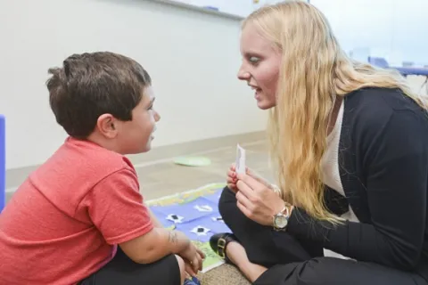 A speech therapist with blonde hair sitting on the ground during a session with a little boy