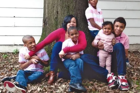 A woman sitting at the base of a tree surrounded by five children wearing pink and red shirts outdoors