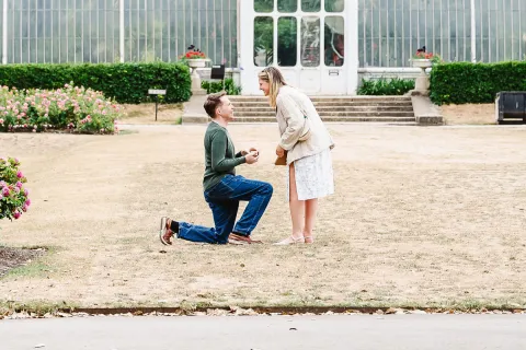 A man kneels with a ring box, proposing to a woman in a garden. They smile at each other in front of a large glass greenhouse