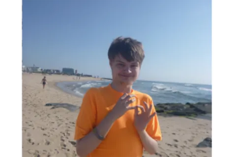 A young man in an orange shirt looks at the camera while standing on a sandy beach 