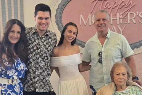 Five family members pose together indoors beneath a “Happy Mother’s Day” sign, beside a wheelchair user.