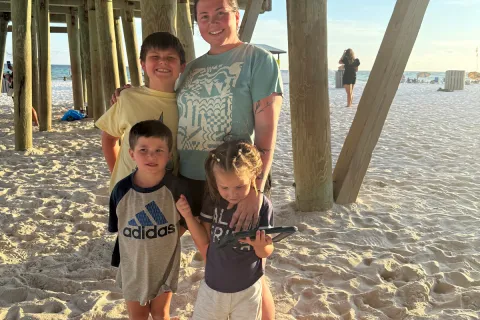 Four people stand together on a sandy beach beneath a wooden pier during late afternoon