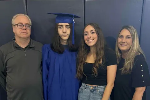 Graduate in blue cap and gown posing with family against a dark blue wall