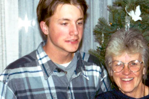 A picture of a young man in a plaid shirt and his mother in a navy shirt in front of a Christmas tree, smiling. 