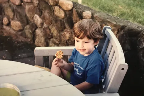 Kerry Magro as a child sitting in a white chair eating a cookie.