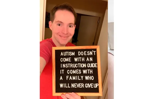 Man in a red shirt holds a letterboard sign that reads "Autism doesn't come with an instruction guide, it comes with a family who will never give up" 