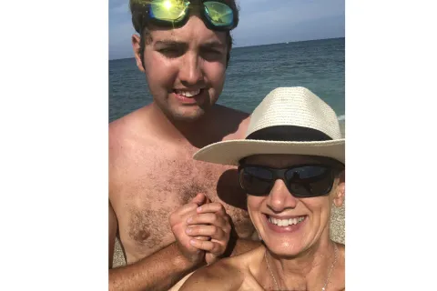 A mother and son are standing in front of the ocean for a selfie. The son has green googles on the top of his head, his mom has a white straw hat and is smiling. They both are smiling