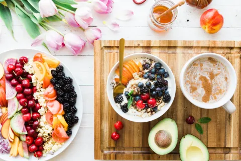 Overhead view of breakfast spread with yogurt bowl, fresh fruit, avocado halves, coffee, honey jar, and tulips on white table