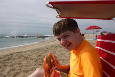 A young boy in an orange shirt sits on the beach under a red umbrella. He is holding a device and smiling at the camera 