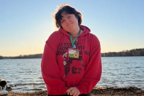 Person in red hoodie standing on sandy lakeshore at sunset, ducks nearby, water and trees in background