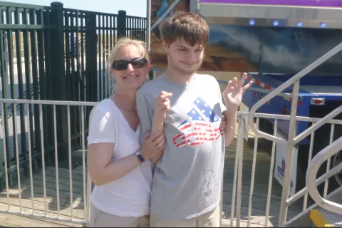 A mother stands behind her teenage son at a outdoor broadwalk setting. She has blonde hair and sunglasses and he is wearing a grey t-shirt with an American flag logo on it 