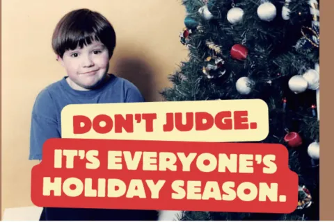 A young boy in a navy shirt sits next to a christmas tree with the words "don't jude. It's everyone's holiday season." 