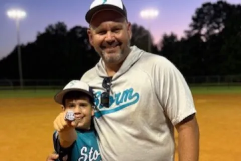 Smiling man and young baseball player posing on a lit field, the boy holding up a ring proudly
