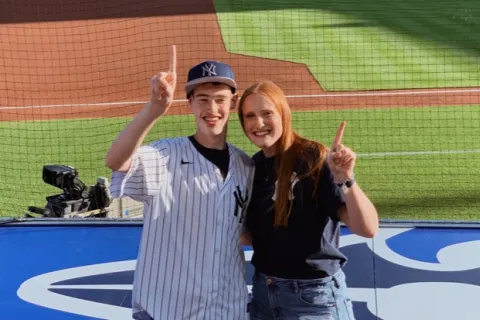 Smiling young man in Yankees jersey and woman posing at baseball stadium holding up number one fingers