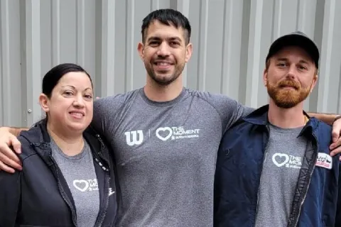 Three Wilson Sporting Goods employees wearing Autism Speaks shirts pose together outdoors in front of a metal building.