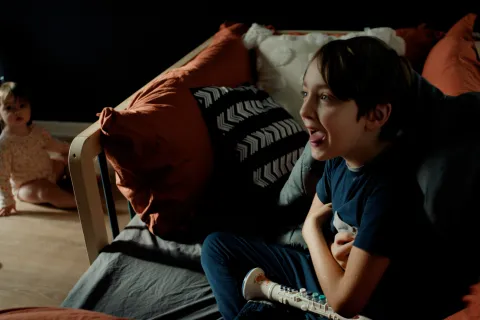 A boy laughs on a orange sofa with his tongue out and is wearing navy colored clothes 