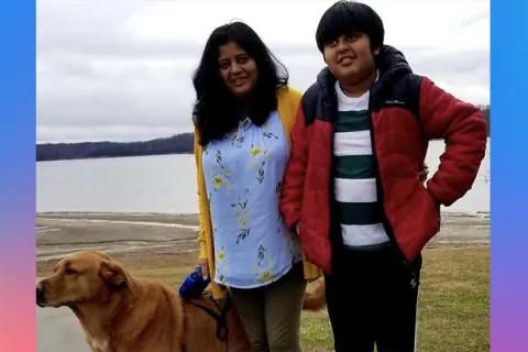 A yellow lab dog stands beside a mother and son who are smiling. The mother is in a yellow  sweater and a blue top and the son has his hands on his hips and has a red puffer jacket on. They are standing in front of a lake on a cloudy day 