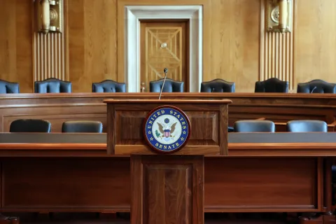 Wooden podium with US Senate seal in front of two lines of long committee desks with black chairs