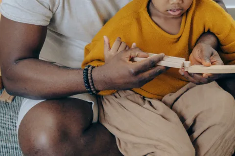 child wearing a yellow sweater and sitting on his dads lap playing with blocks