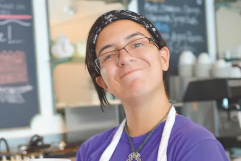 Photo of young woman in an apron smiling in a coffee shop.