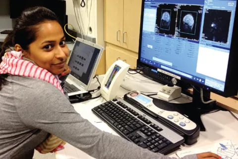 Woman sitting at a desk across from a computer. The computer has images of brain scans.