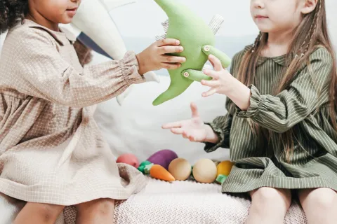 two young girls taking turns playing with a green dinosaur toy