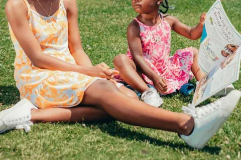 two Black children sitting in the grass and looking at a book