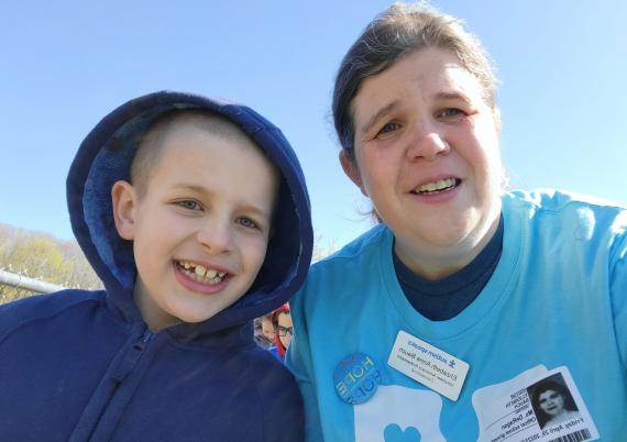 A mom and a son smile at the camera in blue autism shirts on a sunny day 